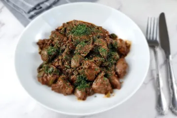 A plate of keto chicken liver, garnished with herbs, accompanied by a fork and knife, and a bowl of sauce on a marble surface.