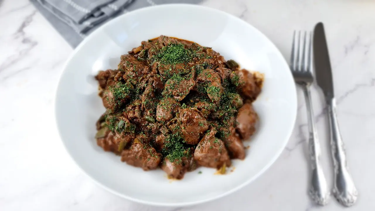 A plate of keto chicken liver, garnished with herbs, accompanied by a fork and knife, and a bowl of sauce on a marble surface.