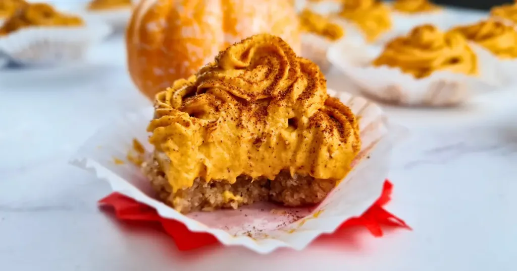 A photo of 1 keto pumpkin spice cheesecake bites arranged on a table, with some whipped cream and cinnamon on top. The photo has a light white background and a small pumpkin on the side. The photo looks appetizing and festive