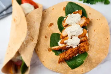 A photo of a keto chicken wrap with ranch dressing on a marble countertop. The wrap is made of a light brown tortilla filled with chicken, spinach, and a white sauce. The chicken is cooked and seasoned, the spinach is fresh and green, and the white sauce is creamy and tangy. There is a bunch of parsley and a gray and white striped towel in the background.