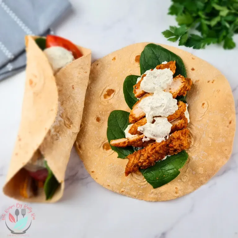 A photo of a keto chicken wrap with ranch dressing on a marble countertop. The wrap is made of a light brown tortilla filled with chicken, spinach, and a white sauce. The chicken is cooked and seasoned, the spinach is fresh and green, and the white sauce is creamy and tangy. There is a bunch of parsley and a gray and white striped towel in the background.