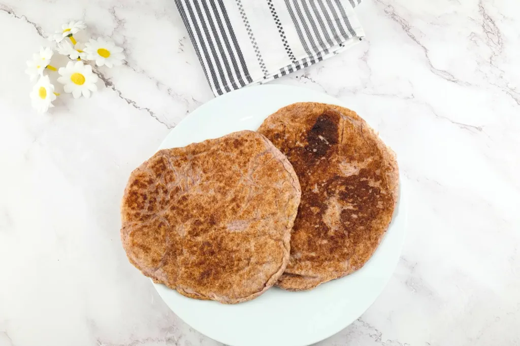 A stack of four round and thin tortillas made with almond flour and psyllium husk on a white plate. The tortillas are low-carb, gluten-free, vegan, and paleo-friendly. They have a golden brown color and a slightly cracked surface. The plate is on a wooden table with a blue napkin and a fork. The image has a text overlay that says “2 Ingredient Keto Tortillas with Almond Flour: Vegan & Paleo”