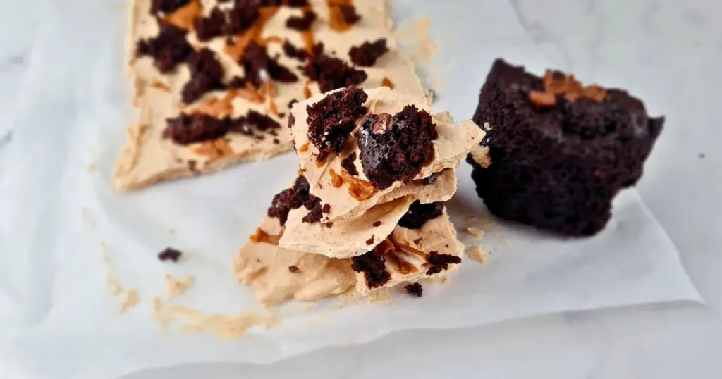 This is a photo of a frozen yoghurt bark with peanut butter and brownie. The bark is rectangular in shape and has a light brown color. It is topped with crumbled brownie and chocolate chips. The bark is cut into smaller pieces and is on a white parchment paper on a white marble countertop.
