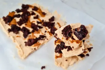 Frozen yoghurt bark with peanut butter and brownie. It is rectangular in shape and has a light brown color. It is topped with crumbled brownie and chocolate chips. The bark is cut into smaller pieces and is on a white parchment paper on a white marble countertop.