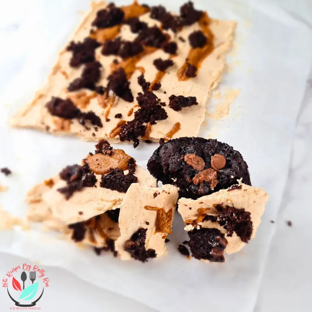 This is a photo of a frozen yoghurt bark with peanut butter and brownie. The bark is rectangular in shape and has a light brown color. It is topped with crumbled brownie and chocolate chips. The bark is cut into smaller pieces and is on a white parchment paper on a white marble countertop.