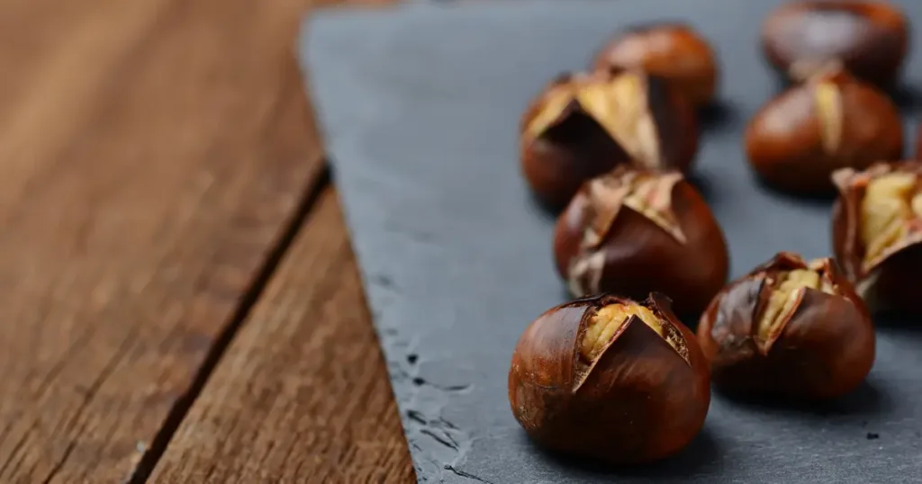 A photo of a wooden bowl of peeled water chestnuts on a wooden surface with a knife. The water chestnuts are white and round. The bowl is a dark brown color and appears to be made of wood. The knife is a silver color with a black handle. The background is a light blue color.