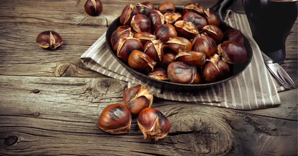 A photo of a bowl of roasted water chestnuts on a wooden table with a glass of dark beer. The water chestnuts are dark brown and have been roasted until they are slightly charred. The bowl is black and has a white stripe around the rim. The table is made of dark wood and has a rustic texture. There is a glass of dark beer on the table next to the bowl. The background is blurred and consists of a wooden wall.