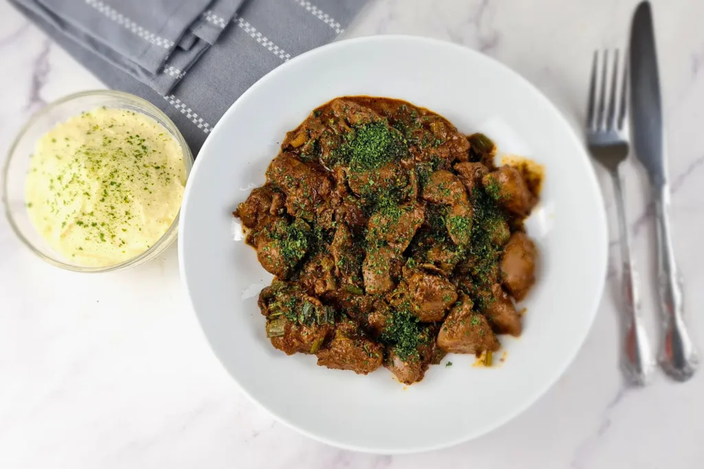 A plate of keto chicken liver with cauliflower mash, garnished with herbs, accompanied by a fork and knife, and a bowl of sauce on a marble surface.
