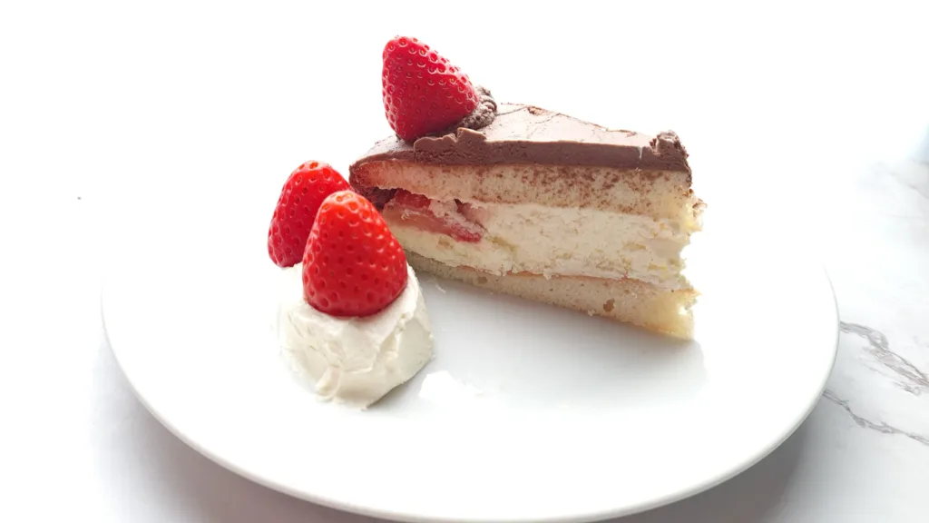 A slice of keto chocolate cake topped with strawberries and a dollop of cream, served on a white plate.