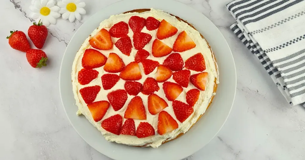 A delicious keto chocolate cake topped with a layer of cream and adorned with sliced strawberries arranged in a circular pattern, presented on a light blue plate. The cake is surrounded by fresh strawberries, white daisies, and a striped cloth on a marble surface.