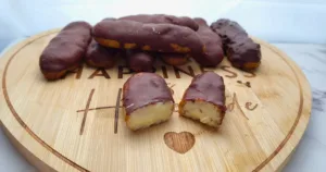 A selection of Keto Chocolate Eclairs displayed on a round wooden board that has “HAPPINESS Homemade” engraved on it. Two eclairs are cut in half, exposing the creamy filling.
