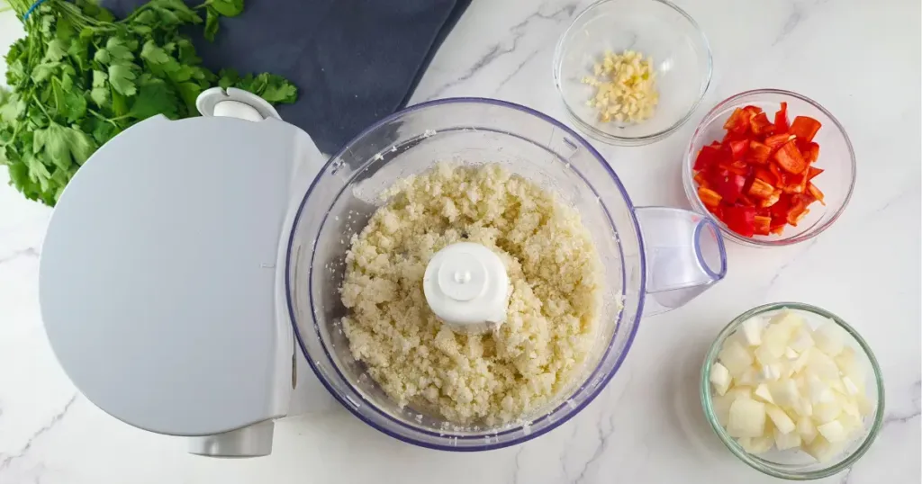 A photo of a light blue food processor and four small glass bowls with chopped cauliflower, red bell pepper, onion, garlic, and cilantro on a white marble countertop. The photo shows the ingredients for making cauliflower rice, a low-carb and keto-friendly alternative to regular rice.