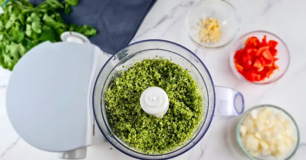 A photo of a light blue food processor and four small glass bowls with chopped broccoli, red bell pepper, onion, garlic, and cilantro on a white marble countertop. The photo shows the ingredients for making broccoli rice, a low-carb and keto-friendly alternative to regular rice.