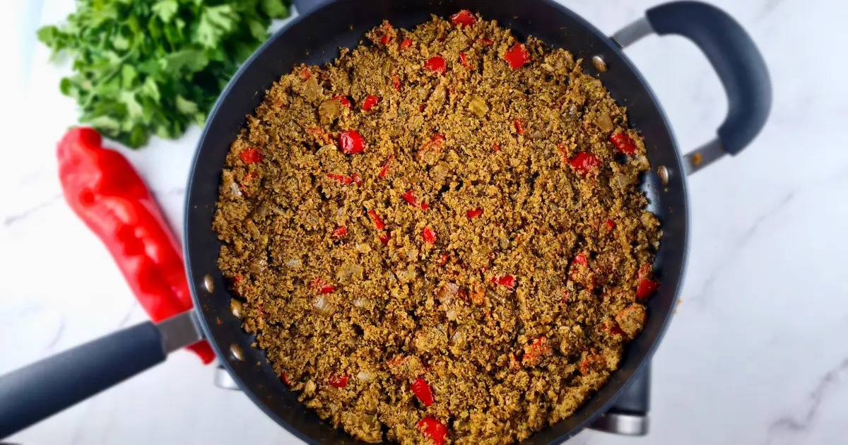 A photo of a black pan filled with a colorful and appetizing dish of Keto Mediterranean Cauliflower & Broccoli Rice, sprinkled with red pepper and onions. The dish is made with cauliflower and broccoli rice, onion, garlic, olive oil, basil, thyme, salt, pepper. It is a low-carb, vegan, and paleo recipe that can be enjoyed as a side dish or a main course. A red pepper is on the left side of the pan, adding some spice and contrast to the dish. The pan is on a white marble countertop, creating a clean and elegant background.