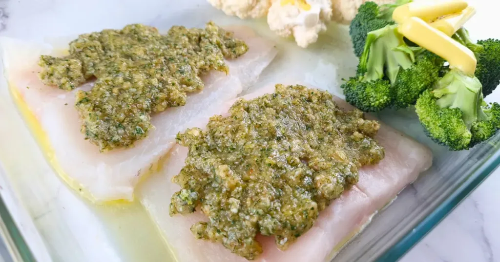 Raw cod fillets topped with a green herb walnut sauce, accompanied by broccoli and cauliflower in a glass dish.