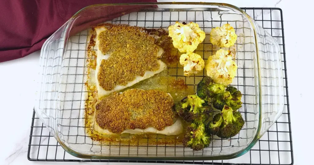 A glass baking dish containing two pieces of Mediterranean cod with a walnut crust, accompanied by roasted cauliflower and broccoli, placed on a wire rack.