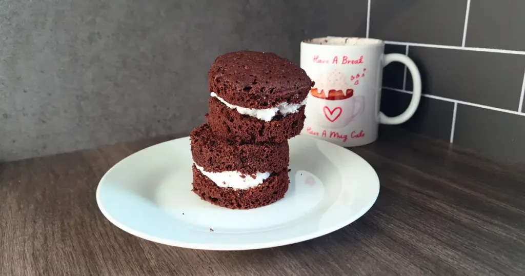Two Keto Oreo Mug Cake with white keto frosting filling, stacked on a white plate, next to a mug with ‘Have A Break Have A Mug Cake’ written on it, against a grey and white tiled background.