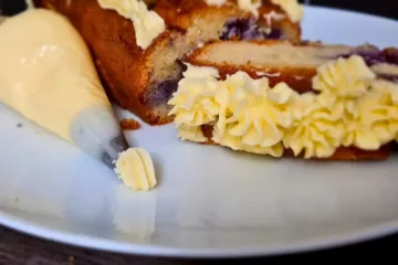 A slice of cake with layers of purple filling and yellow keto royal icing buttercream frosting, next to a piping bag on a white plate.