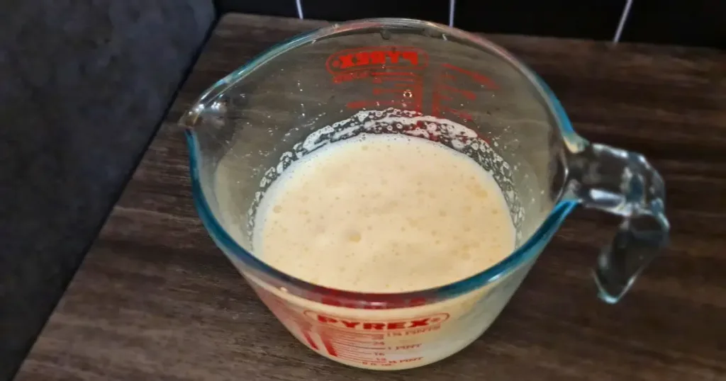 A photo of a glass measuring cup filled with keto Yorkshire pudding batter on a wooden surface. The measuring cup is a Pyrex brand and has red measurement markings on the side. The batter is a pale yellow color and has bubbles on the surface. The measuring cup is on a wooden surface. The background is blurred and dark.