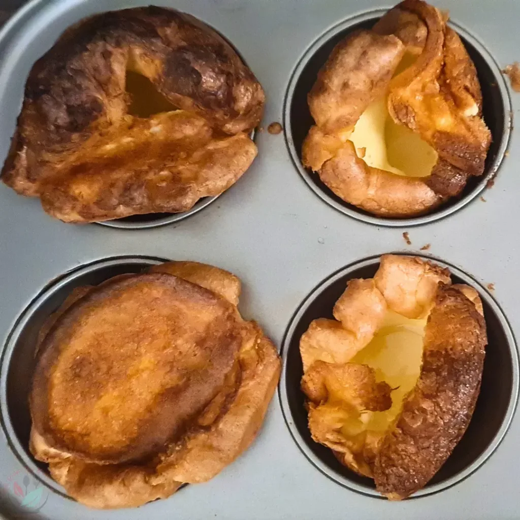 Four keto Yorkshire puddings in a muffin tin. The puddings are golden brown and puffy. The puddings are in a silver muffin tin with four cups. The background is a white countertop.