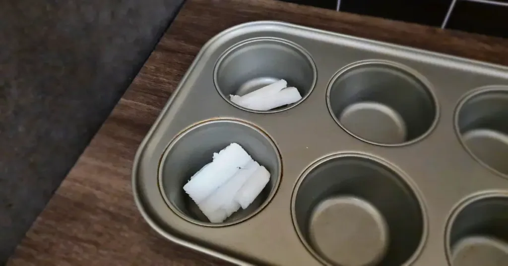 A close up of a keto Yorkshire pudding in a muffin tin with beef dripping in it. The muffin tin is gray and has six cups, but only one pudding is visible. The background is a wooden surface with some crumbs and a fork.