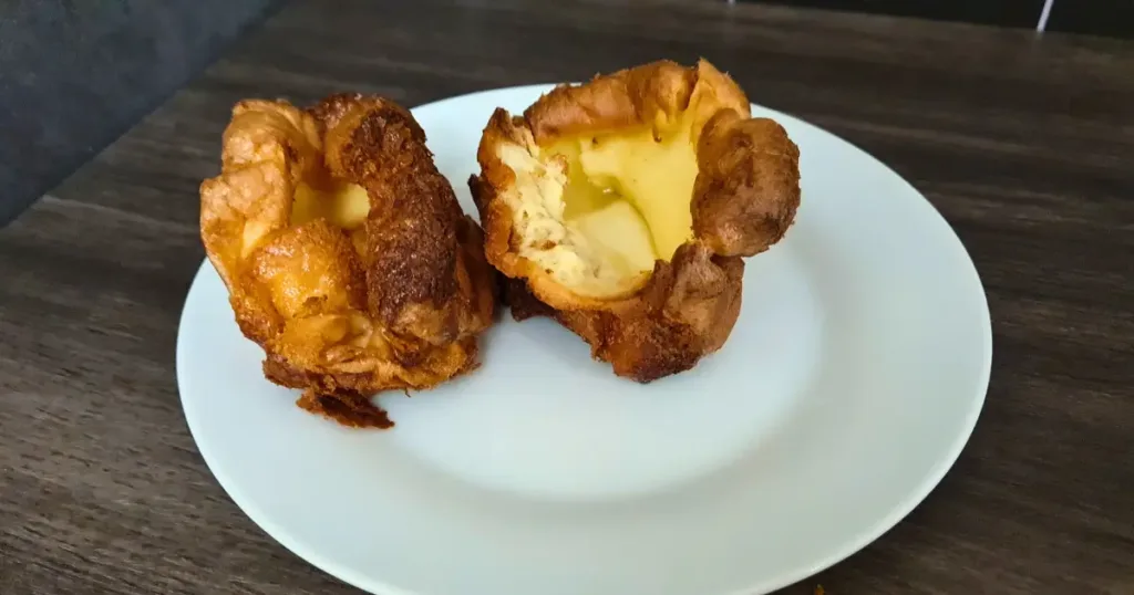 Two keto Yorkshire puddings on a white plate. The puddings are golden brown in color and appear to be freshly baked. The plate is round and white in color. The background is a dark wooden surface.