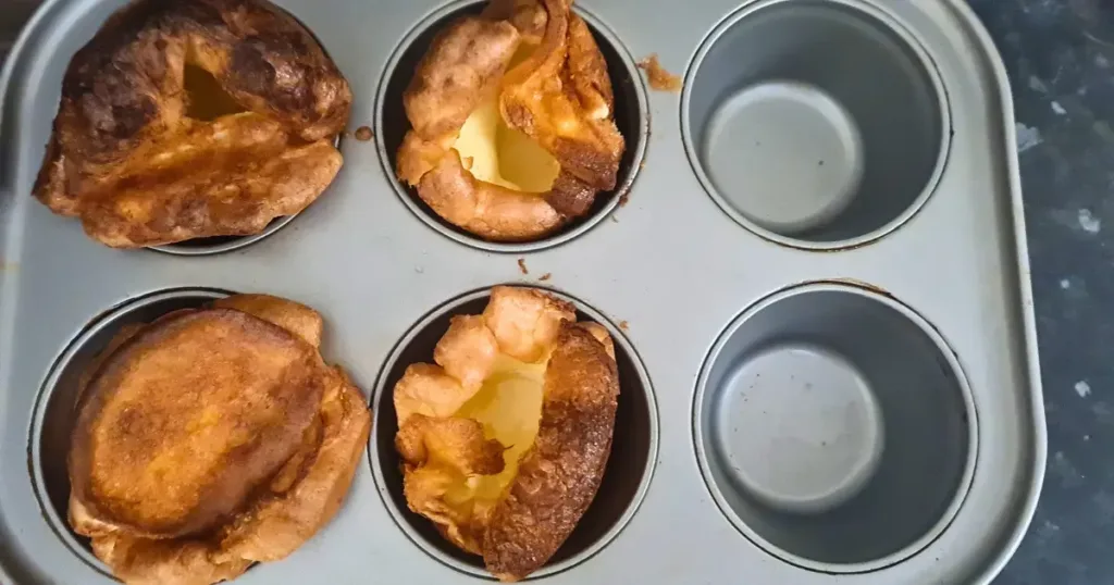 A close up of a keto Yorkshire pudding in a muffin tin. The pudding is golden brown and has a crispy crust and a soft center. The muffin tin is gray and has six cups, but only four puddings are visible. The background is a wooden surface with some crumbs and a fork.