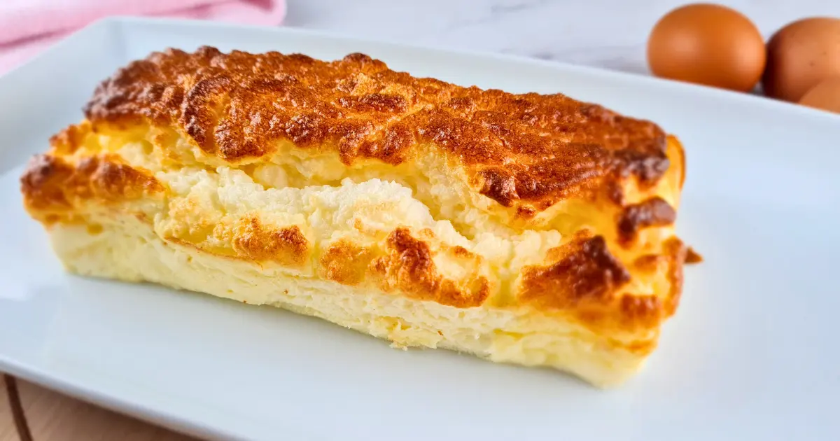 A golden brown, fluffy piece of 2 Ingredient Cottage Cheese Bread served on a white plate, with a pink cloth and three eggs in the background.