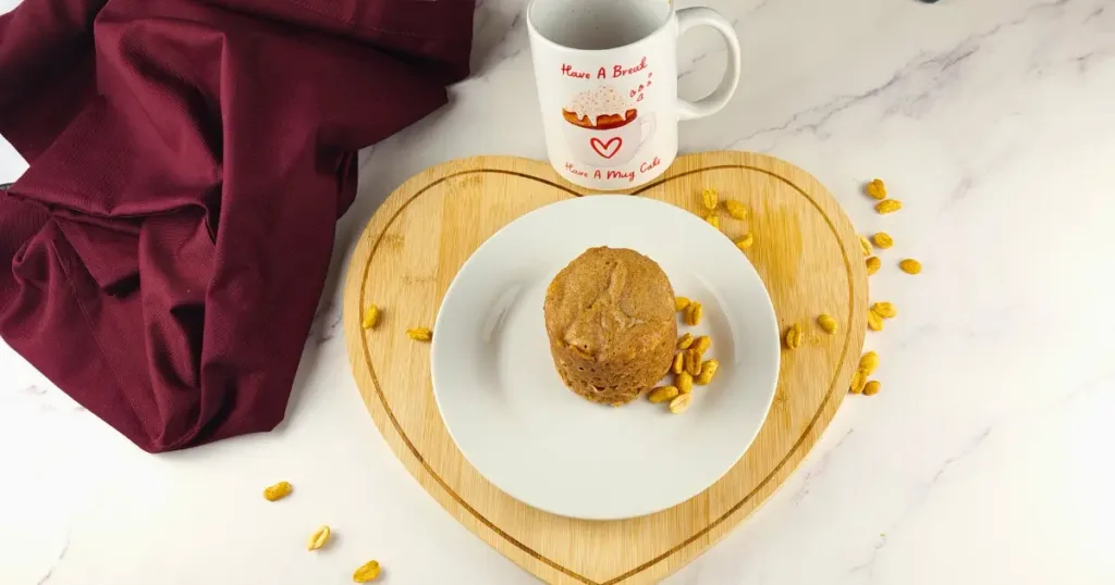 A delicious flourless keto peanut butter mug cake sits on a white plate, surrounded by scattered peanuts. The plate is on a heart-shaped wooden board next to a mug with ‘Have A Break Have A Mug Cake’ written on it, all set against a marble countertop.