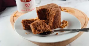 A moist, freshly baked flourless keto peanut butter mug cake on a white plate, with a piece on a fork ready to be enjoyed. A mug with ‘Have A Mug Cake’ written on it and scattered peanuts are in the background.