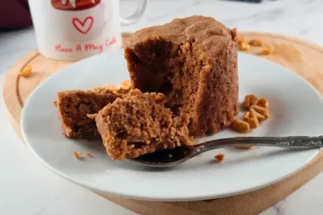 A moist, freshly baked flourless keto peanut butter mug cake on a white plate, with a piece on a fork ready to be enjoyed. A mug with ‘Have A Mug Cake’ written on it and scattered peanuts are in the background.
