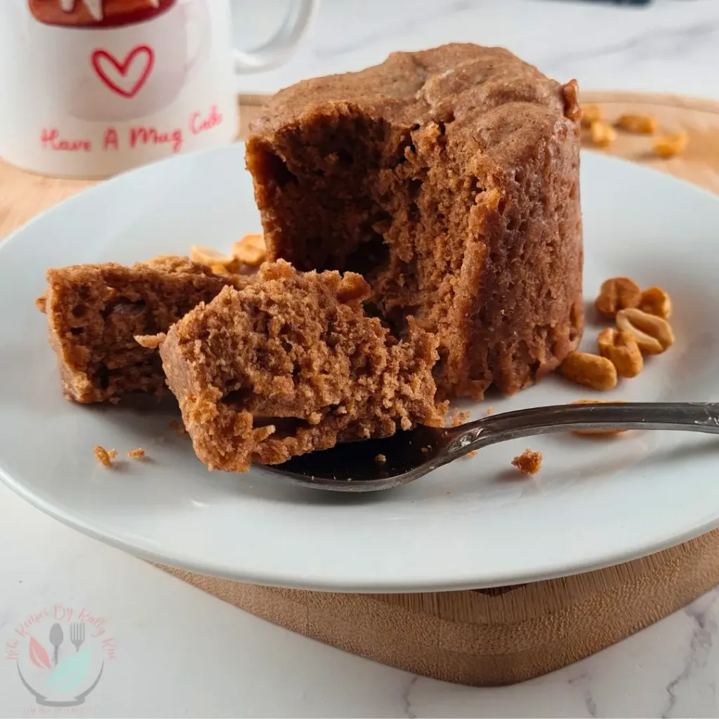 A moist, flourless keto peanut butter mug cake on a white plate, with a piece on a spoon ready to be enjoyed. A mug with ‘Have A Mug Cake’ written on it is partially visible in the background.