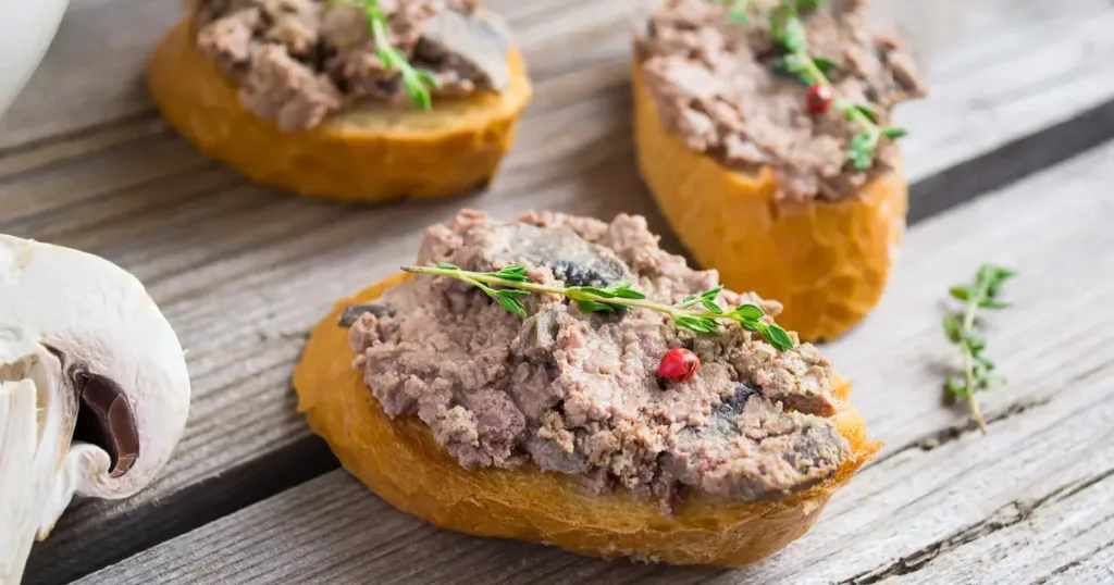 Slices of bread topped with beef liver pate, garnished with herbs and spices, placed next to a mushroom on a wooden surface.