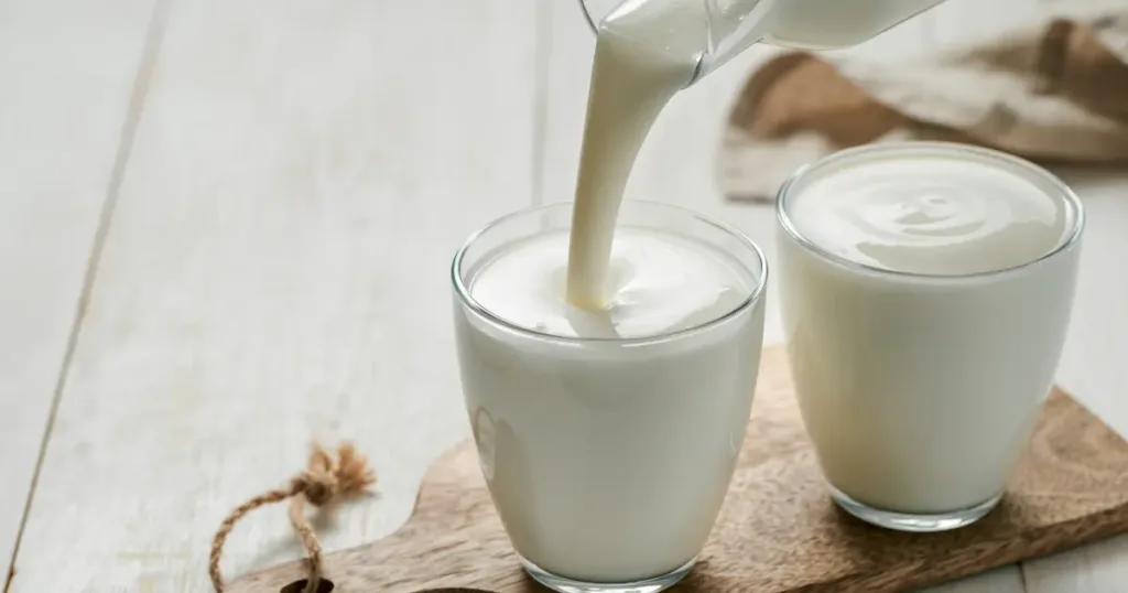 Buttermilk being poured into a clear glass, with another full glass beside it, both on a wooden board.