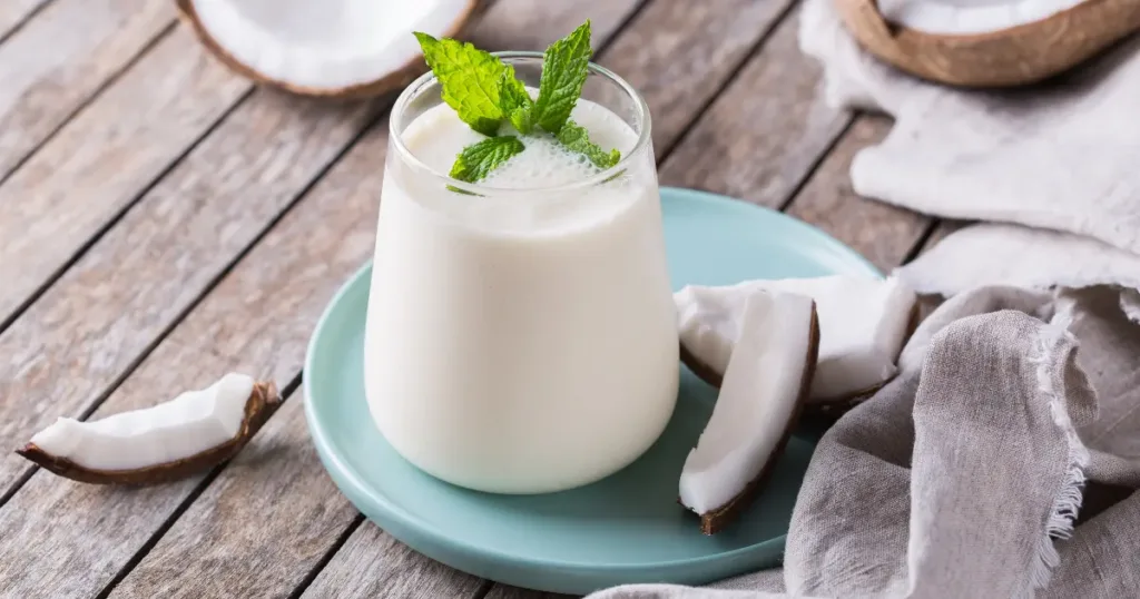 A refreshing glass of buttermilk garnished with fresh mint leaves, served on a teal plate, surrounded by coconut pieces and a cloth on a wooden surface.