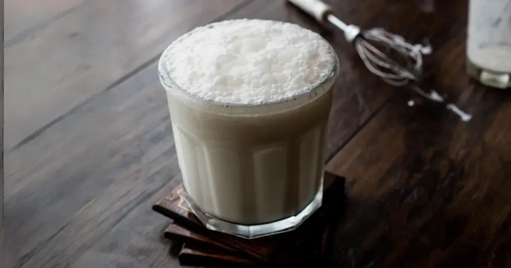 A frothy glass of buttermilk topped with whipped cream, placed on a dark wooden surface beside cinnamon sticks and a whisk.