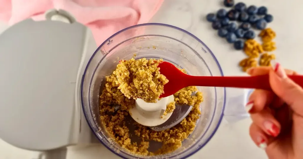 A hand holding a red spatula with a scoop of cheesecake walnut crust mixture, taken from a food processor bowl. Fresh blueberries and walnuts are scattered on the white countertop beside it.