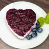 A heart-shaped keto blueberry cheesecake topped with a glossy, rich blueberry layer, served on a white plate with fresh blueberries and a sprig of basil, on a wooden surface.