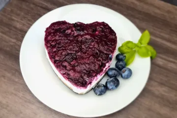 A heart-shaped keto blueberry cheesecake topped with a glossy, rich blueberry layer, served on a white plate with fresh blueberries and a sprig of basil, on a wooden surface.