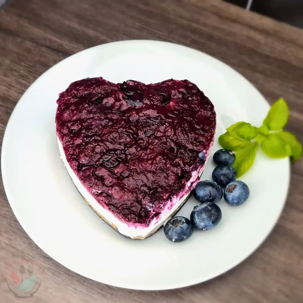 A heart-shaped keto blueberry cheesecake garnished with fresh blueberries and a sprig of basil, served on a white plate.