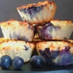 A stack of golden brown Keto blueberry muffins made with coconut flour, garnished with fresh blueberries, presented on a dark round tray.