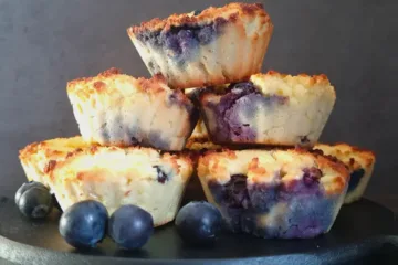A stack of golden brown Keto blueberry muffins made with coconut flour, garnished with fresh blueberries, presented on a dark round tray.
