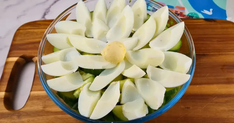 Sliced boiled eggs arranged in a flower pattern on top of green vegetables, placed in a glass bowl on a wooden cutting board.