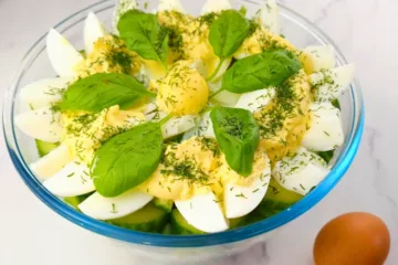 A bowl of keto egg salad with halved boiled eggs, creamy yellow dressing, fresh green basil leaves, and sprinkled herbs on a marbled surface with a whole egg beside it.