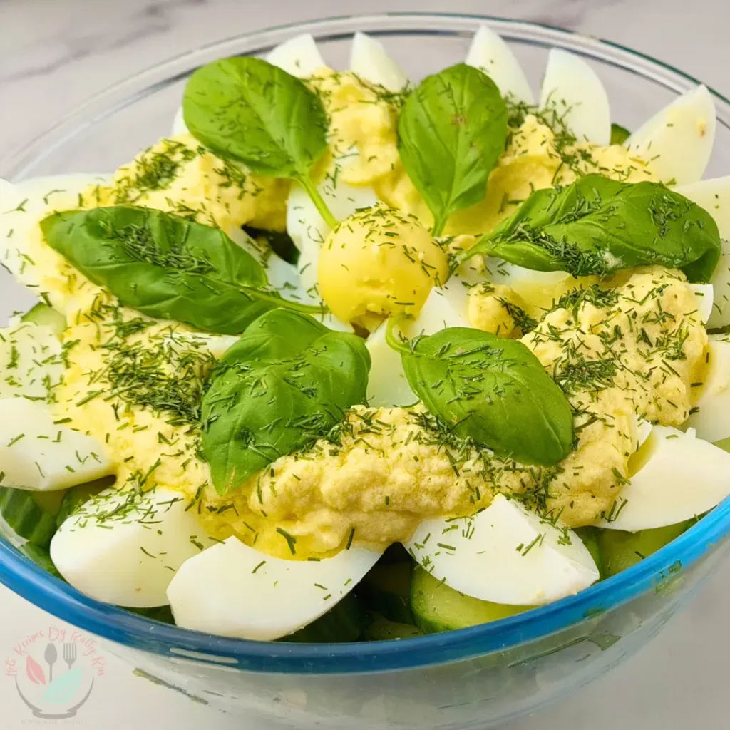 A colorful keto egg salad with boiled eggs, creamy yellow dressing, fresh green basil leaves, and sprinkled herbs in a clear bowl.