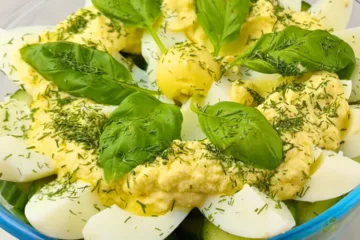 A colorful keto egg salad with boiled eggs, creamy yellow dressing, fresh green basil leaves, and sprinkled herbs in a clear bowl.