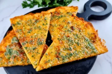 Four slices of golden brown Keto Garlic Bread garnished with herbs, served on a black round cutting board.