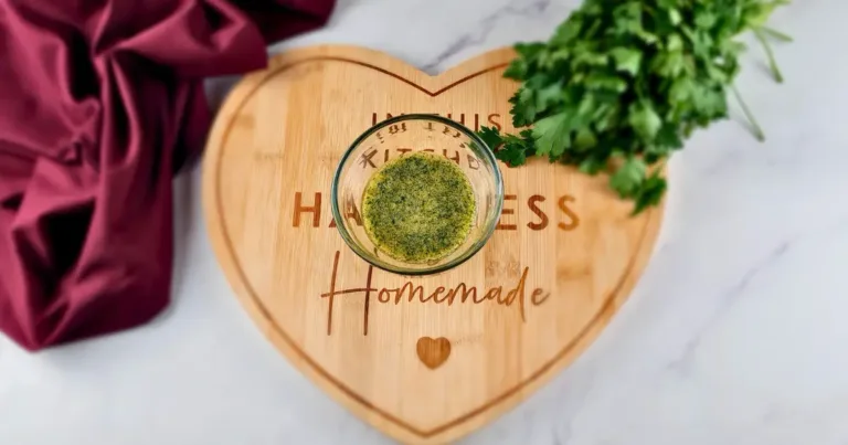 A bowl of Keto Garlic Butter placed on a heart-shaped wooden board that reads ‘Happiness is Homemade’, with a bunch of fresh herbs and a folded burgundy cloth beside it, all set on a marble surface.