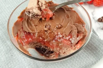 A Keto Mug Cheesecake with chocolate and strawberry jam, fresh strawberries, and chocolate chips on a marble surface.