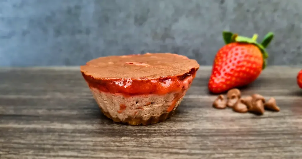 A delectable keto mug cheesecake with a rich chocolate layer on top and a succulent strawberry jam layer in the middle, placed on a wooden surface, accompanied by fresh strawberries and chocolate chips.